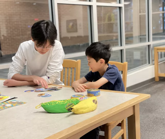 Photo of a young boy sitting beside an older male youth at a table in the Library. On the table are puzzles, books and a game.