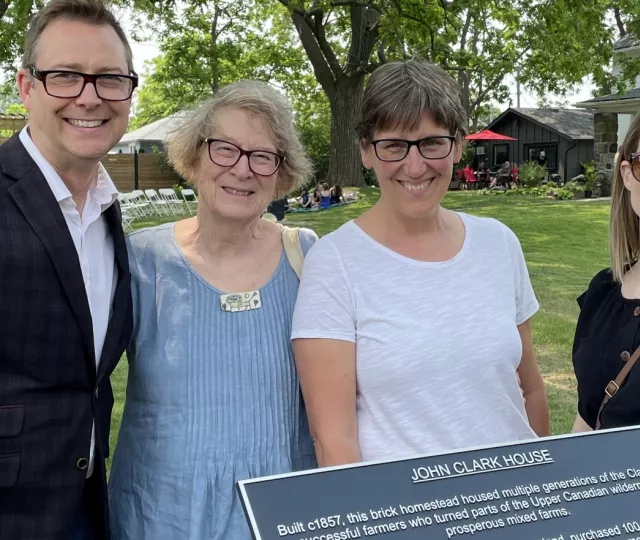 Four people stand behind a plaque commemorating John Clark House. In the background is a white brick house with people gathering on the lawn.