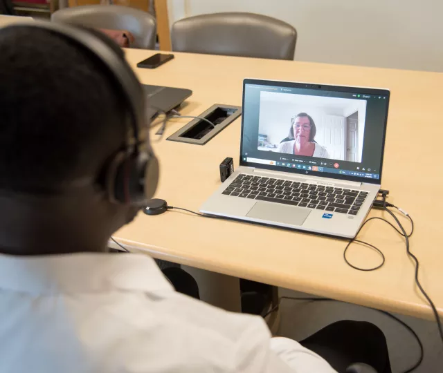 A man with headphones on looking at a laptop screen which is displaying a virtual healthcare appointment.