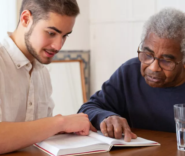 a caregiver and an elderly individual sitting together at the table. The caregiver is assisting the elderly man read.