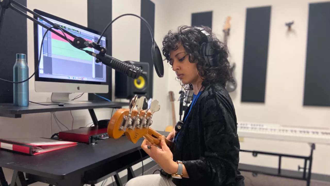 Photo of a musician with headphones on sitting on a stool in the audio recording studio in The Labs and playing guitar. In the background is a screen speakers and a keyboard.