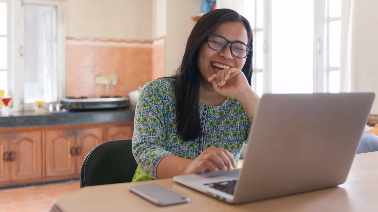 Photo of a woman sitting in a kitchen with a laptop on a table in front of her.  She is smiling and her phone sits beside her on the counter.