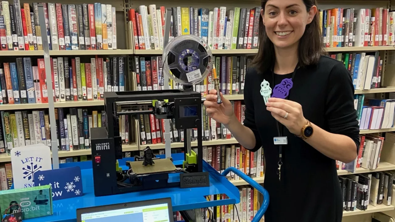 Photo of a librarian standing with a cart on which sits a 3D printer and a laptop. Behind are bookshelves.