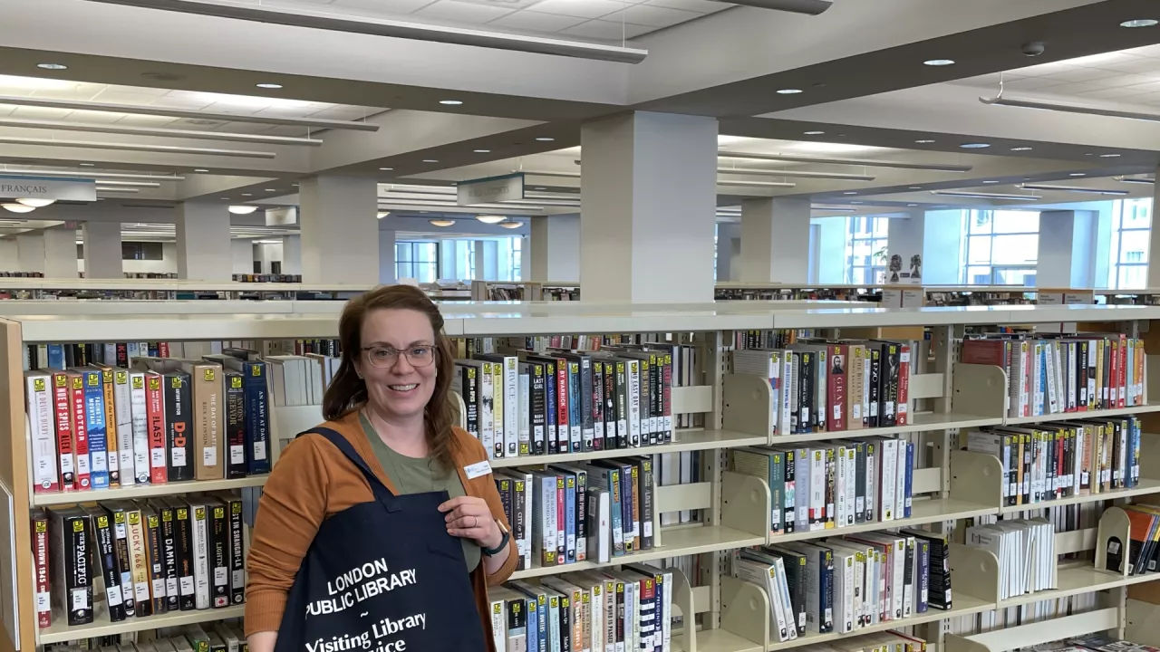 Photo of a librarian holding a Visiting Library Service black bag and standing in front of a book shelf at the Library.