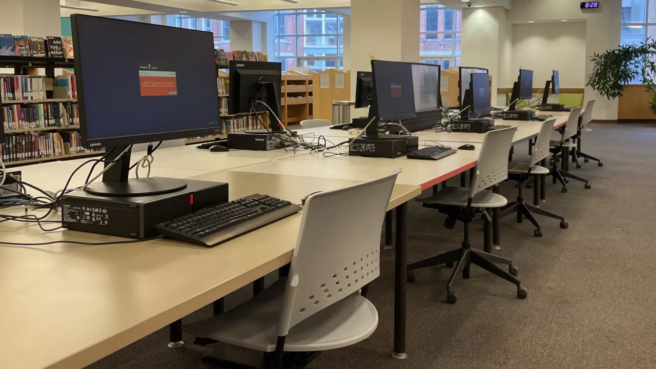 A photo of desktop computers on tables in a line at Central Library. In front of each table is a chair. In the background are bookshelves, windows and a seating area.