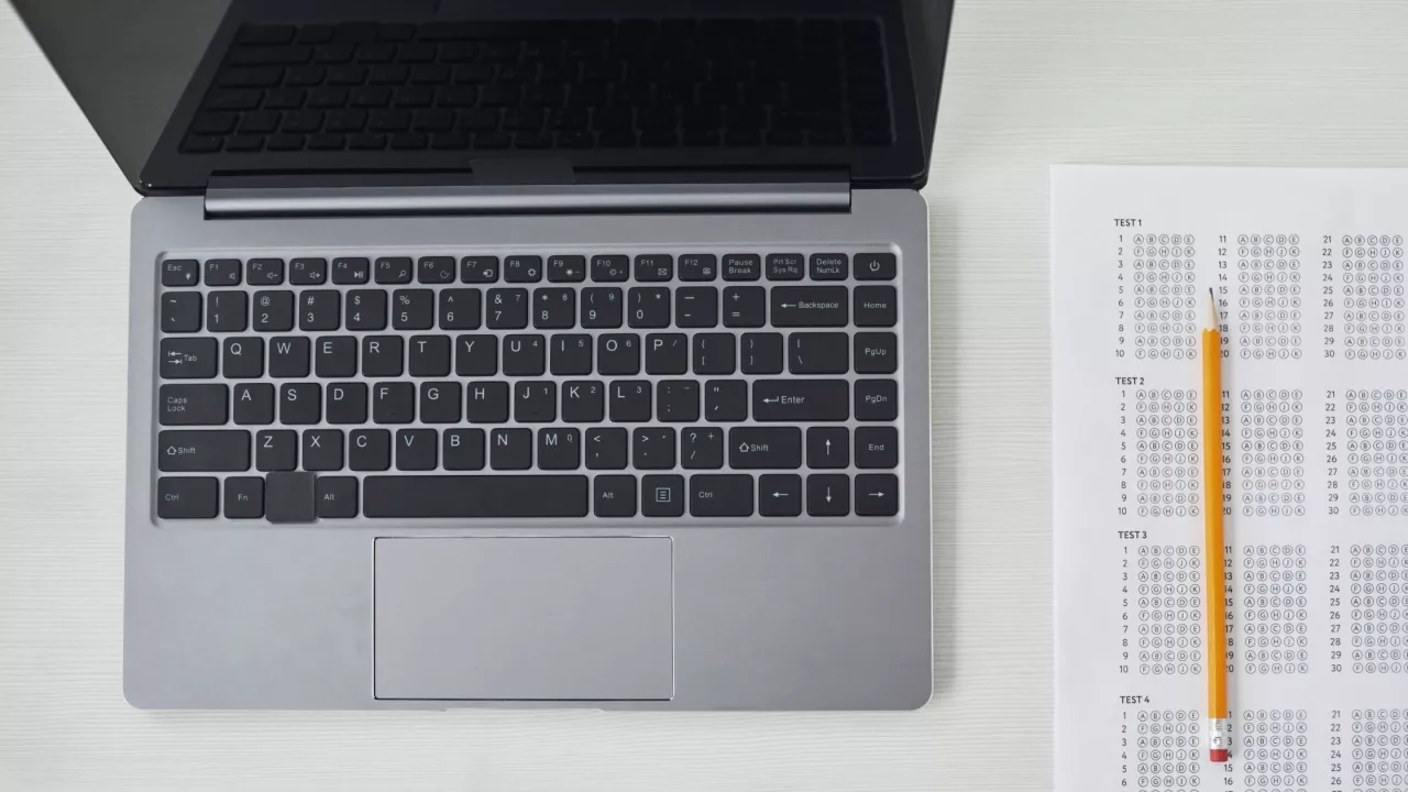 Photo of a laptop on a table. Beside the laptop is a paper with multiple choice questions and a pencil.