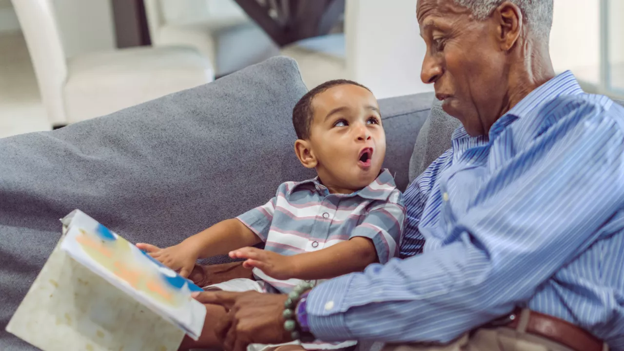 Photo of an older man and young child sitting together on a couch with a picture book. They are looking at each other and are sounding out words. The man holds a picture book and points to it with his finger.