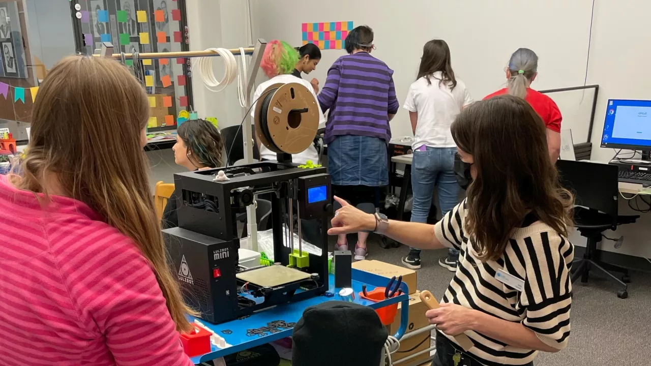 Photo of a group of people in The Labs using the equipment. A library staff member points to a 3D printer in the centre of the room.