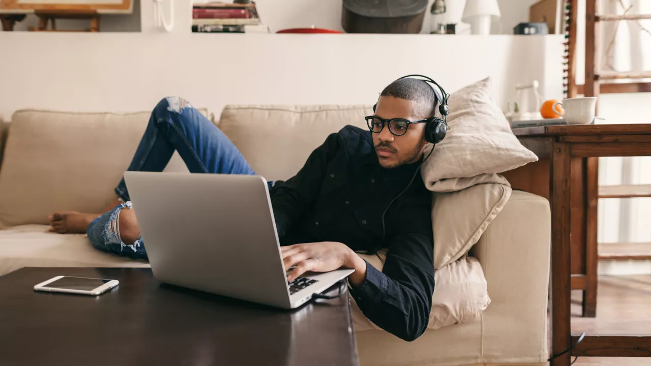 Photo of young man lying on couch and using a laptop that is sitting on a coffee table.  He is wearing headphones and his phone sits on the table.