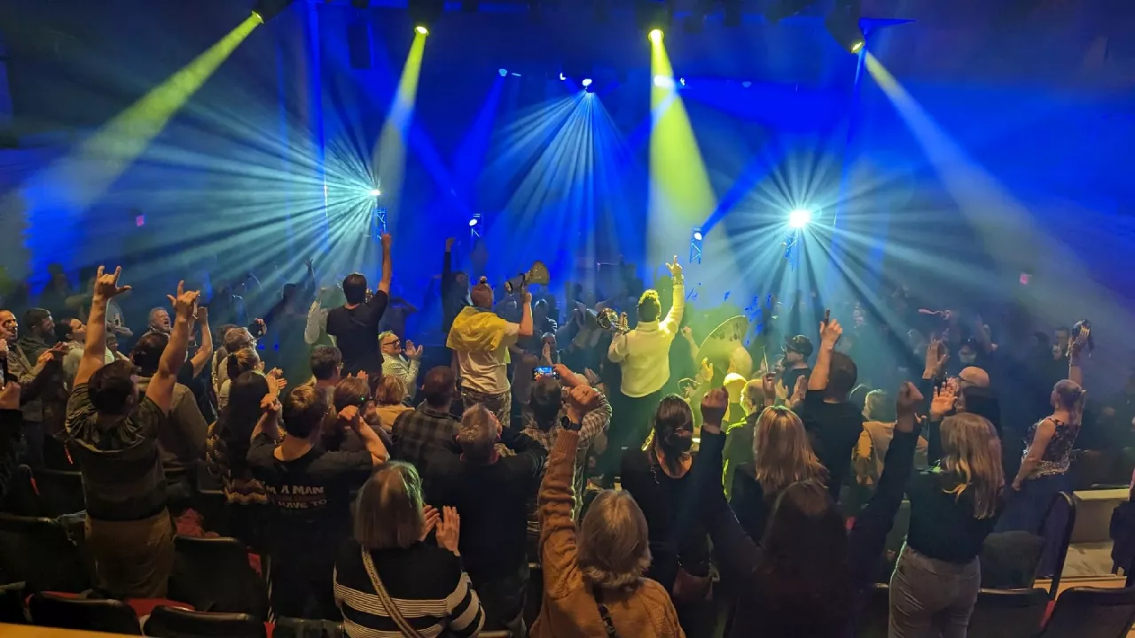 Photo of the backs of a full house at Wolf Performance Hall. The audience is standing and waving their arms in the air. Blue and yellow lights shine brightly from the stage into the audience.ising their arms up