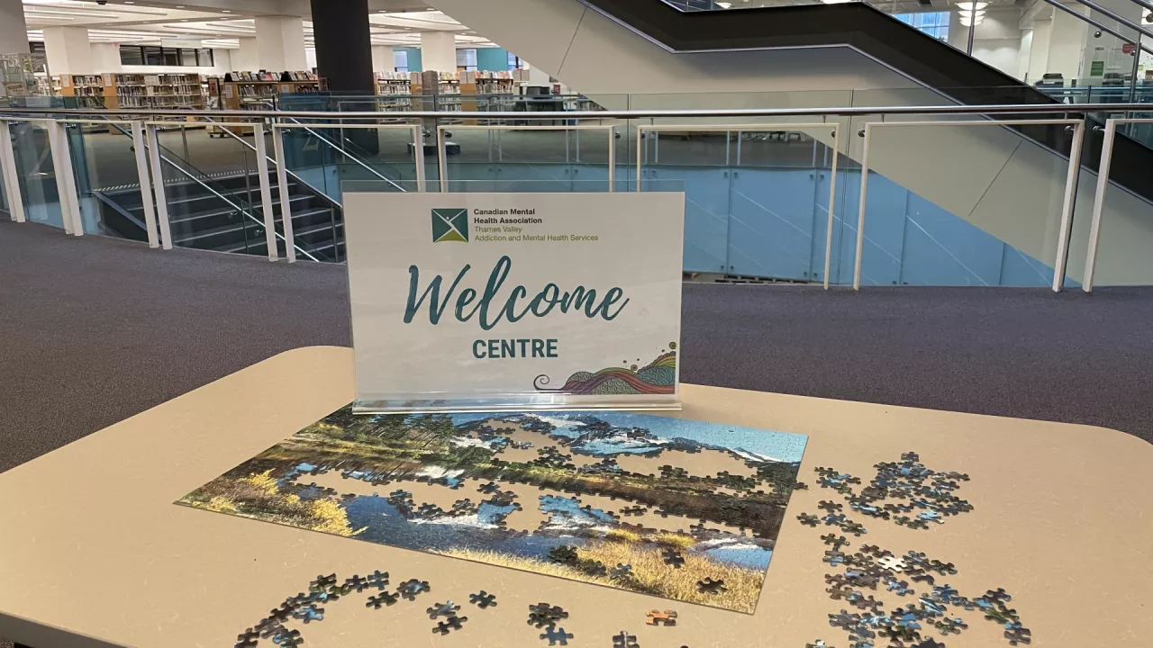 Photo of the Welcome Centre sign sitting on a table in the library on which are scattered puzzle pieces. The poster says Welcome Centre, Canadian Mental Health Association Thames Valley Addition and Mental Health Services