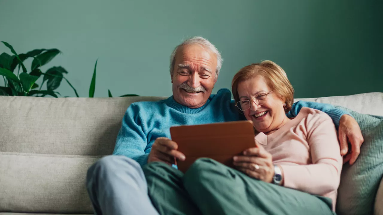 Photo of an older man and woman sitting together on a couch and holding a tablet between them. They are smiling.