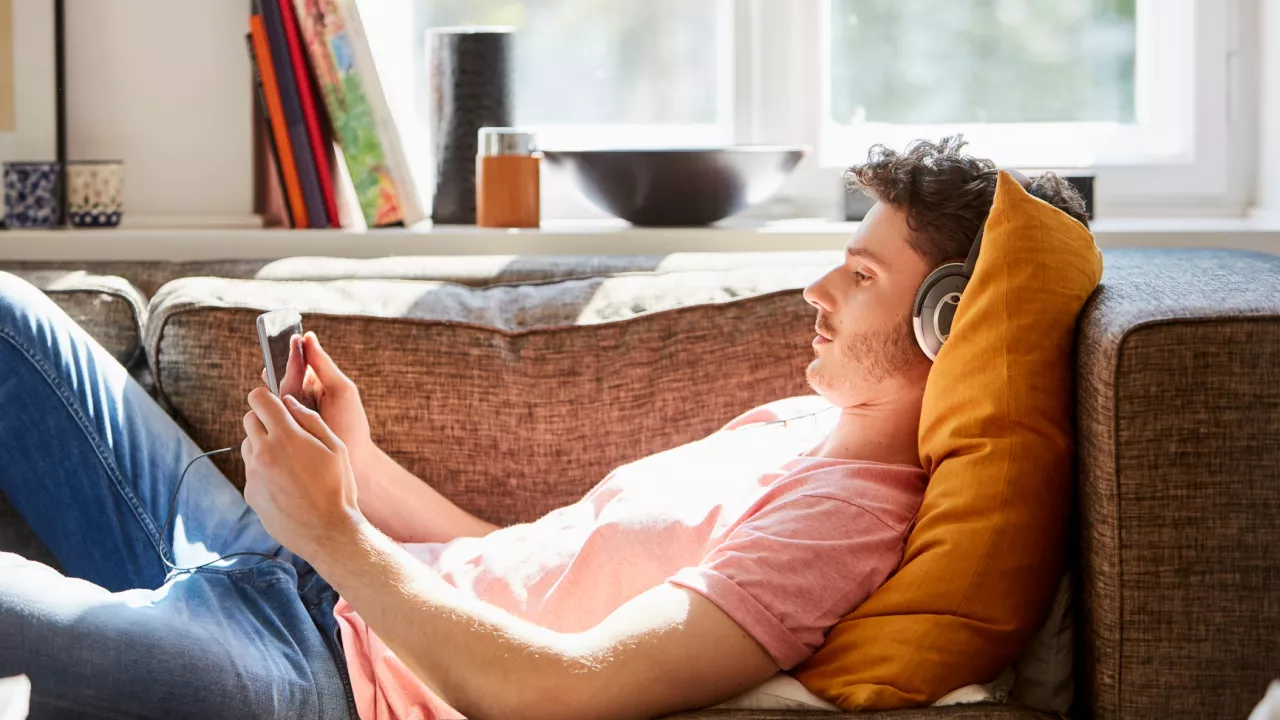 Photo of a young man lying on a couch and holding a tablet. He is wearing headphones and behind him are books on a shelf.