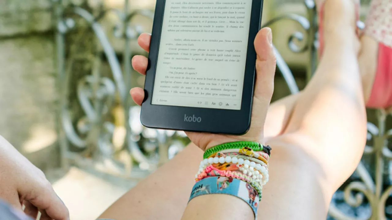 Adult with tattoos and bracelets holding a kobo digital reader while outdoors reading on a patio.