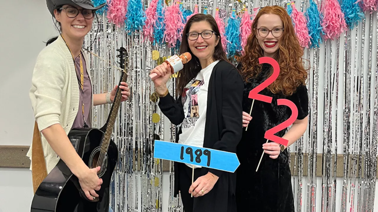 3 women, one with a guitar, one with a microphone and one holding the numbers 22 stand in front of a silver backdrop