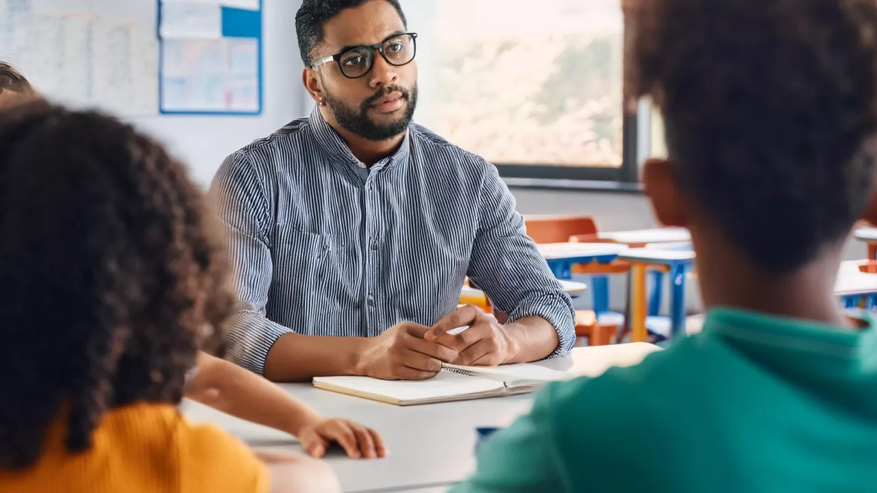 Teacher sitting at desk talking to students in a classroom