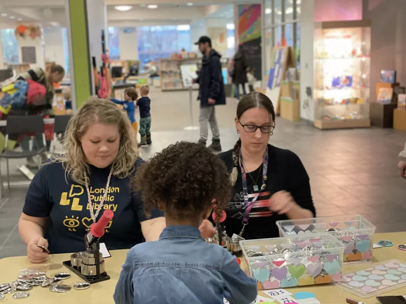 A photo of two library staff sitting at at table at a busy event and making buttons as a young girl joins them.  In the background are families.