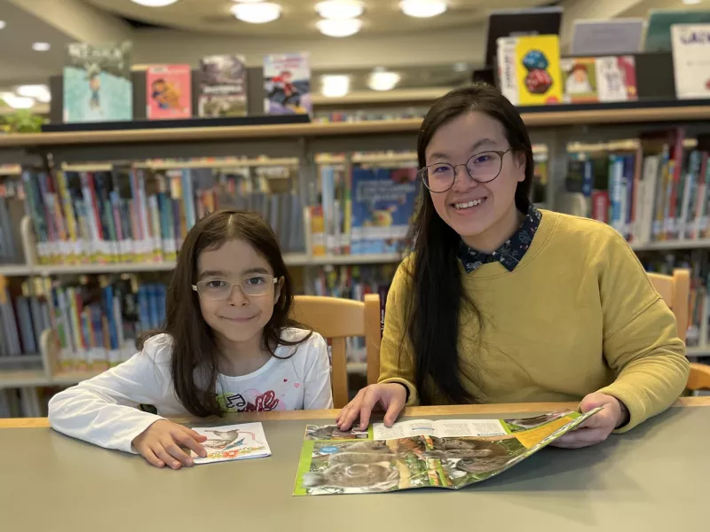 Photo of a child and young woman sitting beside each other at a table and looking up and smiling. They are holding books. In the background are bookshelves.