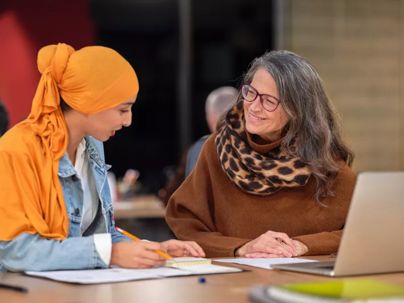 A photo of a younger woman sitting beside another woman in front of whom is a laptop. They are smiling and talking.