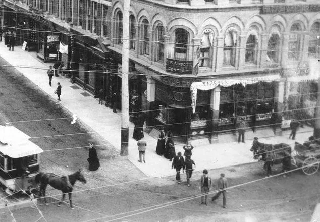 Black and white photograph looking downwards southeast to a building on the corner. Streets are busy with people and horses