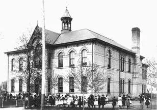 Black and white photo of of a schoolhouse, children gathered on the grounds infront